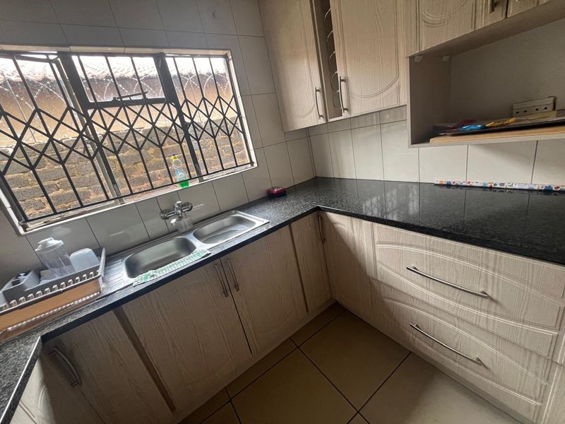 A functional kitchen featuring light wood-grain cabinetry, dark granite countertops, a double basin sink, and a tiled backsplash with security-barred window.