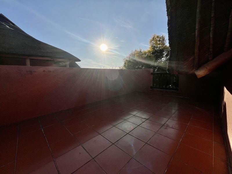 An open-air balcony featuring tiled flooring, a masonry parapet wall, and a traditional thatch roof overhang, offering a bright outdoor space.