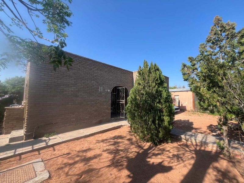 Front exterior view of a single-story exposed brick house featuring an arched entrance with a security gate and surrounding unpaved garden area.