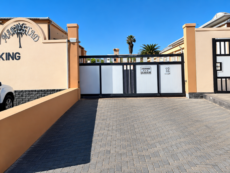 Front entrance view of the apartment complex featuring a paved driveway, secure automated sliding gate, and pedestrian access gate.