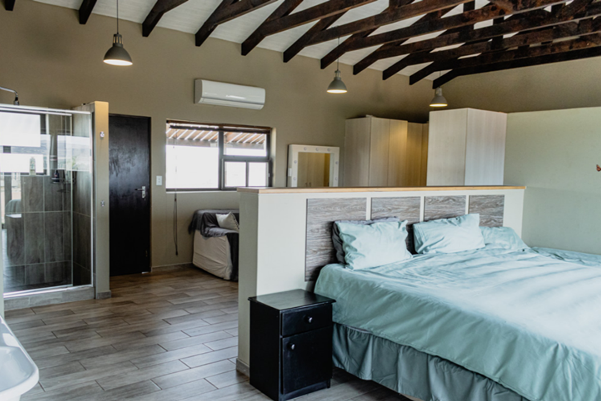 Spacious open-plan bedroom featuring exposed timber roof trusses, tiled flooring, an integrated en-suite shower, and built-in cabinetry.