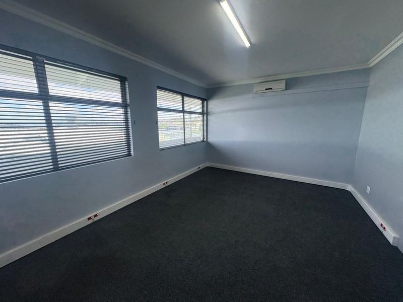 A clean, empty office space featuring carpeted flooring, wall-mounted air conditioning, and large windows with fitted venetian blinds.
