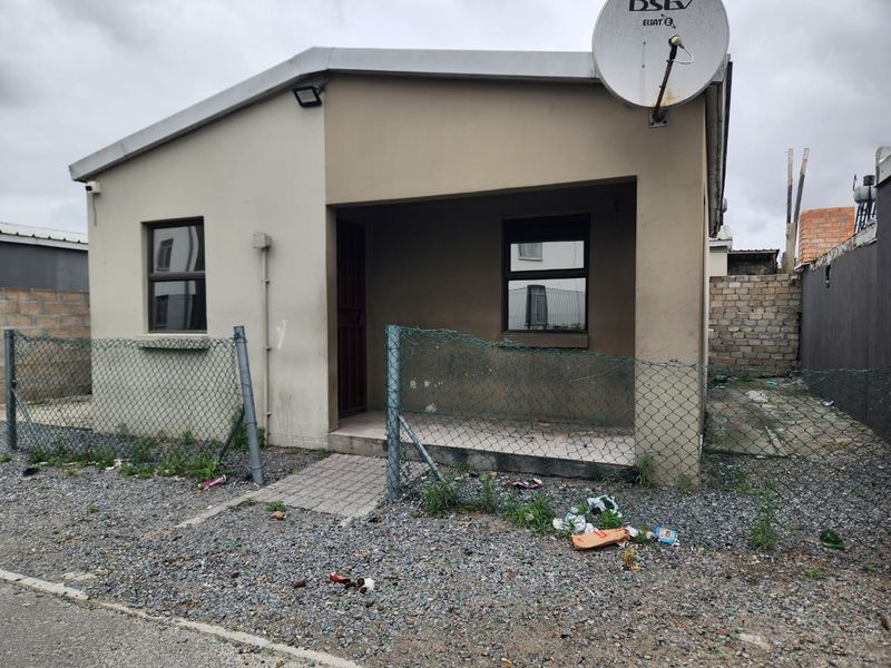 Front elevation of a detached house featuring a small covered entrance, security gate, and satellite dish, set on a plot with damaged perimeter fencing.