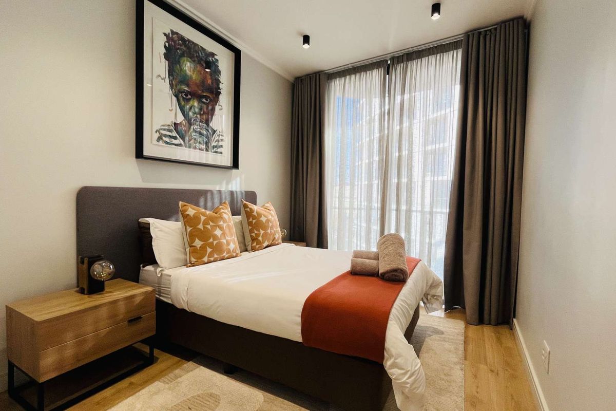 A well-maintained bedroom featuring modern recessed ceiling lighting, light timber flooring, and floor-to-ceiling curtains leading to an outdoor balcony area.