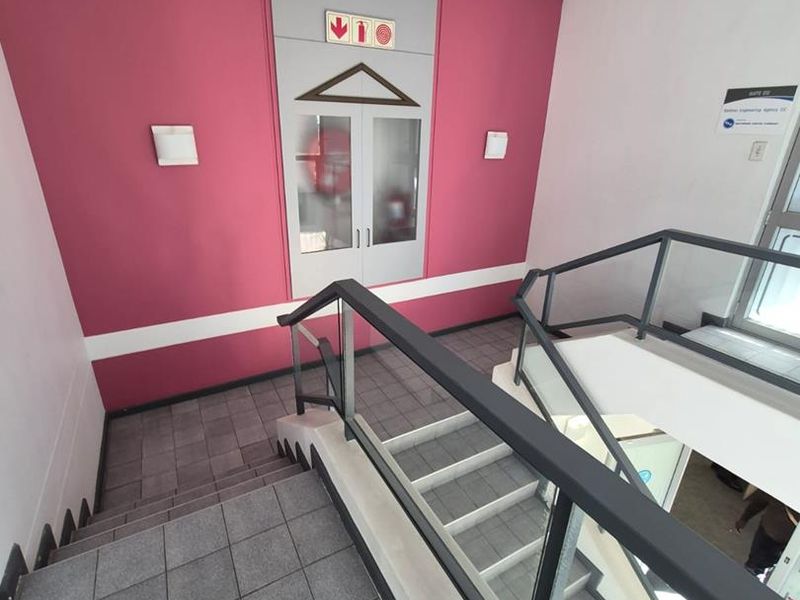 A clean, modern office stairwell featuring tiled flooring, a glass balustrade, and a recessed fire hose cabinet against a vibrant accent wall.