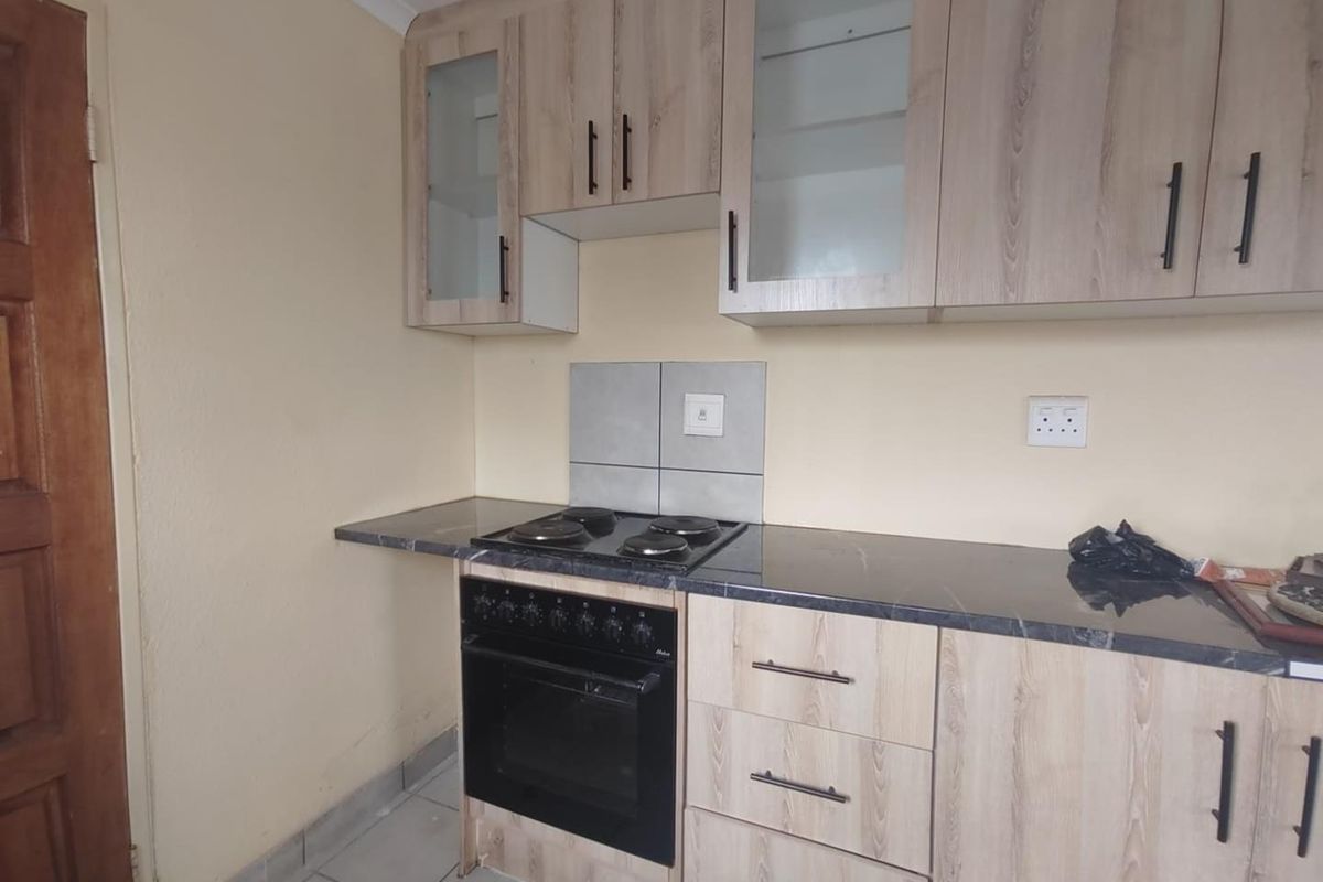 A compact kitchen featuring light wood-finish cabinetry, dark laminate countertops, a built-in electric oven and hob, and neutral tiled flooring.
