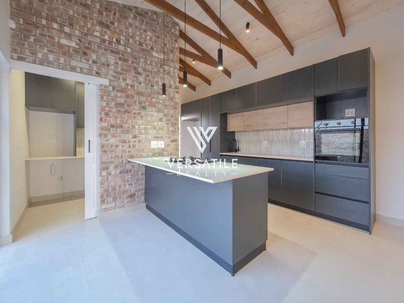 Modern kitchen featuring exposed brickwork, timber ceiling beams, sleek dark cabinetry, a central island, and an adjacent scullery area.