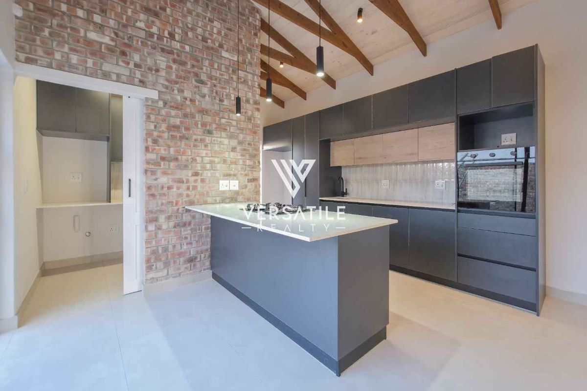 Modern kitchen featuring exposed brickwork, timber ceiling beams, sleek dark cabinetry, a central island, and an adjacent scullery area.