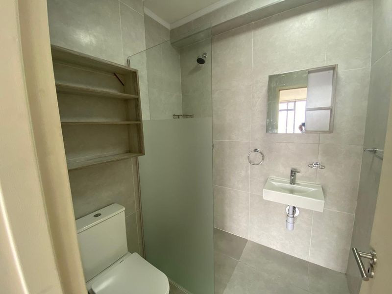 Modern bathroom featuring a wall-mounted basin, glass shower screen, toilet, and built-in shelving with neutral floor-to-ceiling tiling.