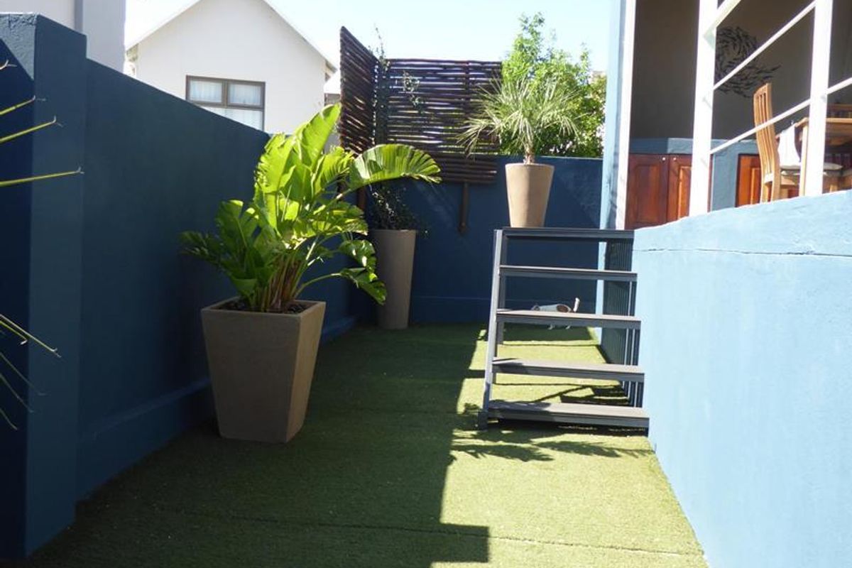 A compact rear courtyard featuring artificial turf, a painted boundary wall, and a metal staircase leading to a covered patio area.