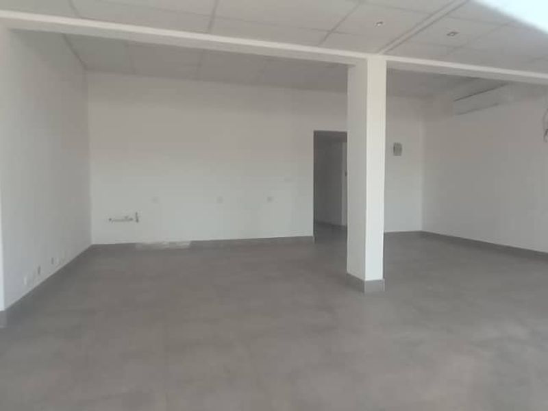 A spacious, empty open-plan retail area featuring neutral tiled flooring, a suspended ceiling with recessed lighting, and a wall-mounted air conditioning unit.