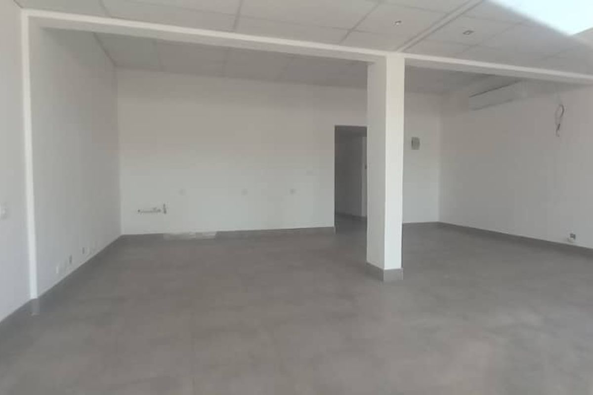 A spacious, empty open-plan retail area featuring neutral tiled flooring, a suspended ceiling with recessed lighting, and a wall-mounted air conditioning unit.