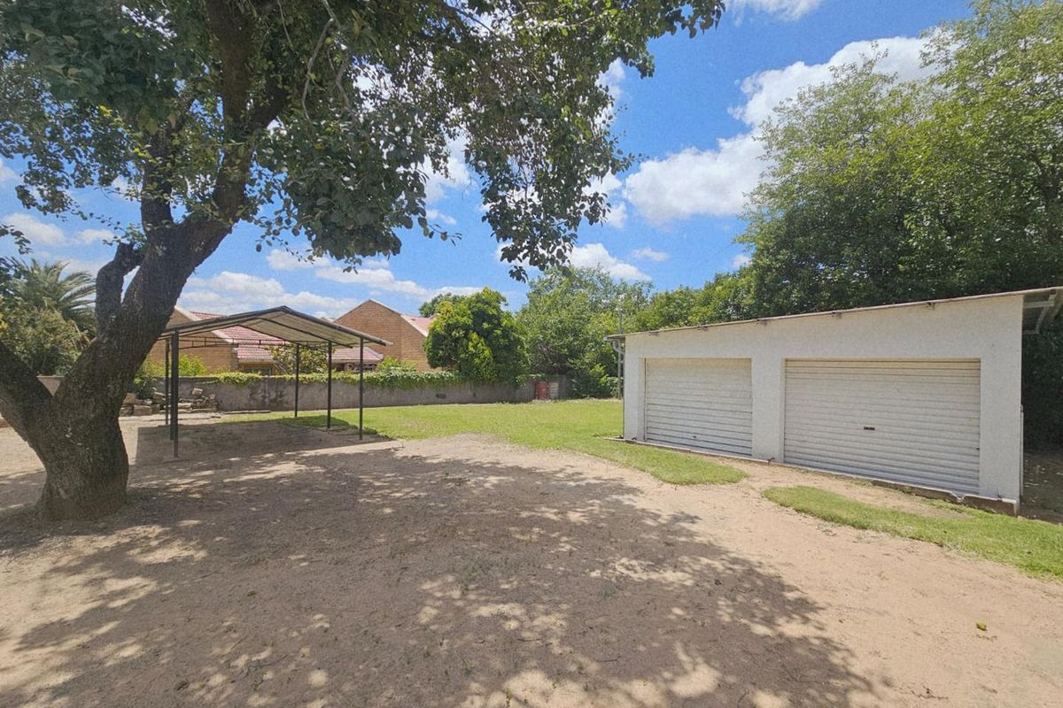 Spacious backyard featuring a double garage, a metal carport structure, and mature trees, offering ample outdoor space and parking potential.