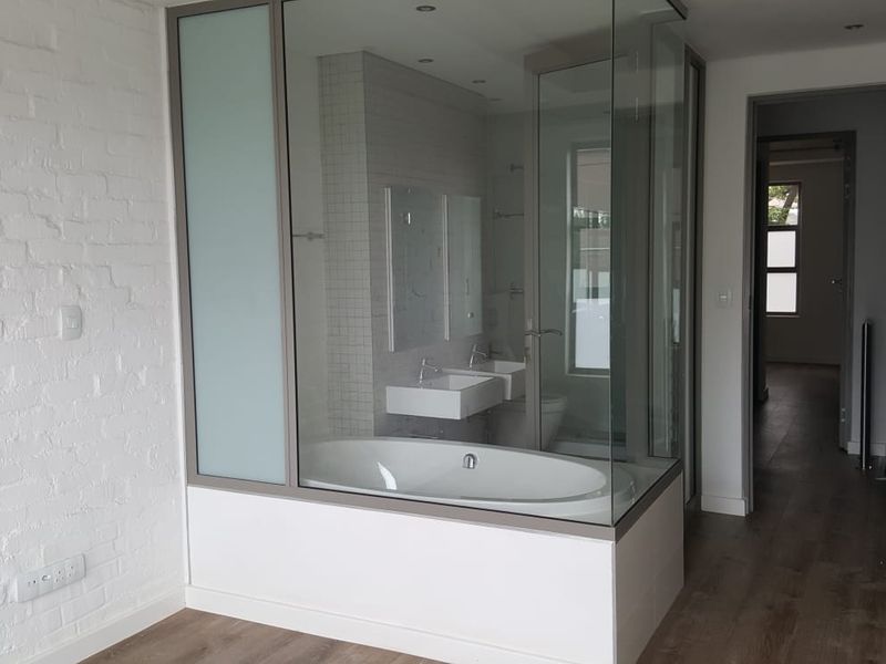 Modern bedroom featuring a unique glass-enclosed en-suite bathroom with a bathtub and double vanity, complemented by an exposed brick wall and built-in storage.