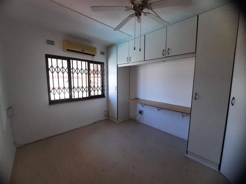 A vacant bedroom featuring built-in cupboards, a desk, a ceiling fan, and a wall-mounted air conditioner with security bars on the window.