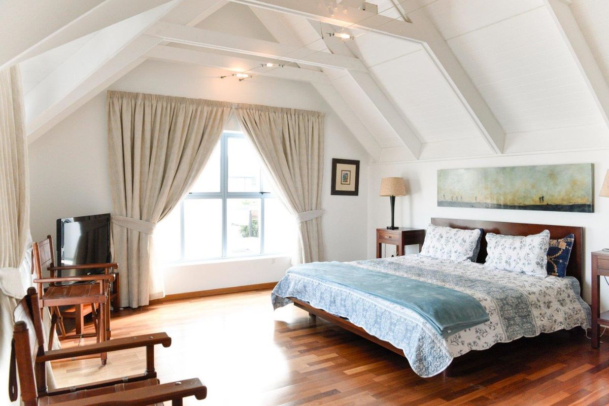 Spacious bedroom featuring vaulted ceilings with exposed timber beams, warm hardwood flooring, and large windows allowing for ample natural light.