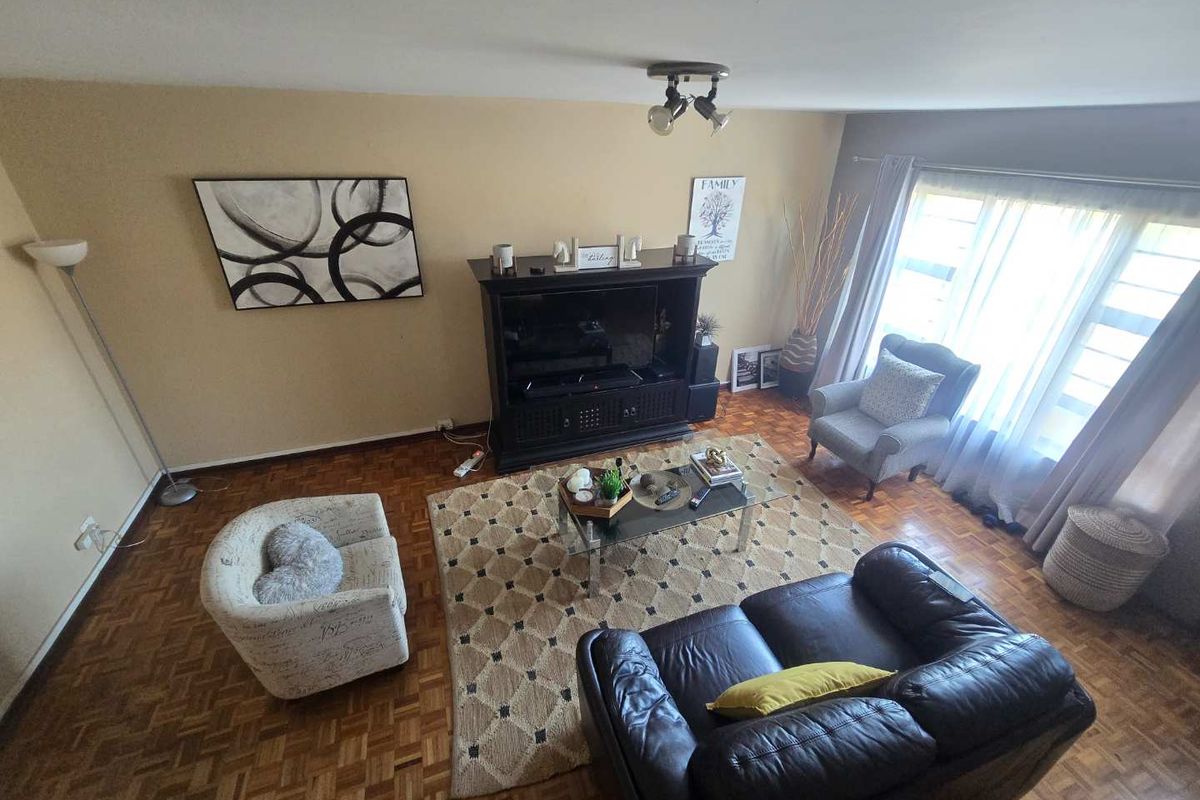 A spacious living room featuring classic parquet flooring, large windows allowing natural light, and a central ceiling-mounted spotlight fixture.