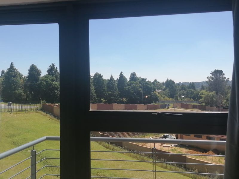 A view from an apartment window looking out over a green lawn, perimeter fencing, and surrounding trees.