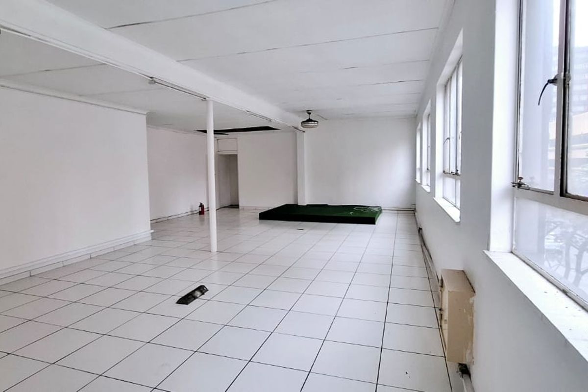 An empty, open-plan office space featuring tiled flooring, large windows providing natural light, and a suspended ceiling structure.