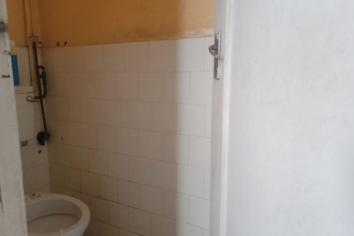 A small bathroom featuring a standard toilet, white wall tiles, and exposed plumbing, requiring cosmetic updates and maintenance.