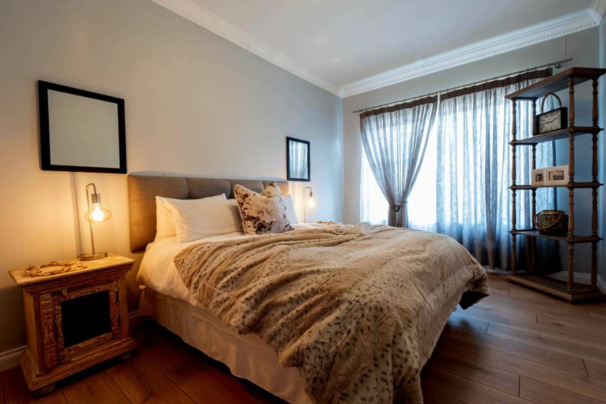A well-maintained bedroom featuring wood-look flooring, crown molding, and large windows with sheer curtains, creating a bright and comfortable living space.
