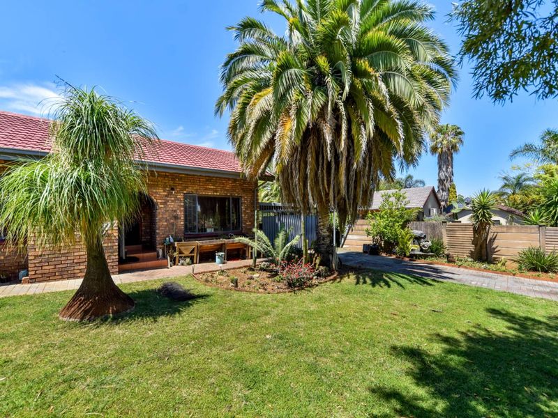 A well-maintained front exterior featuring a classic brick facade, tiled roof, paved driveway, and established garden with mature palm trees.