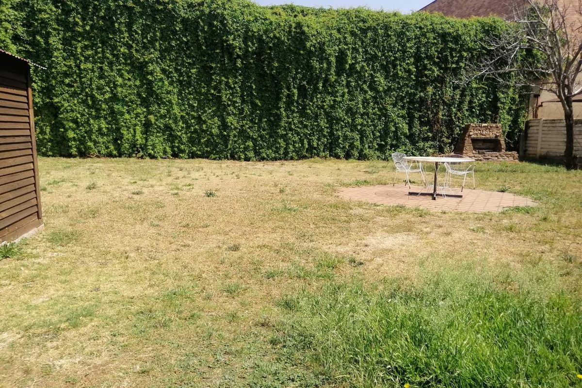 A spacious rear garden featuring a paved circular patio area, a built-in brick braai, and a tall, established ivy-covered boundary wall.
