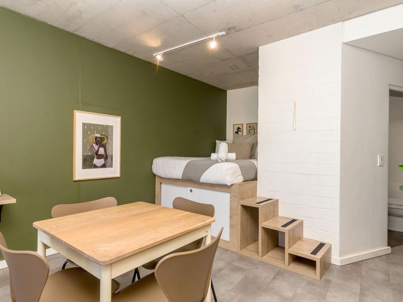 Modern open-plan studio apartment featuring an exposed concrete ceiling, built-in loft bed with integrated storage steps, and a contemporary olive green accent wall.