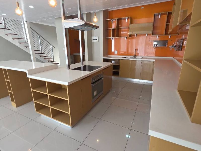 Modern kitchen featuring a central island, built-in oven, electric hob, extractor fan, and sleek cabinetry with orange glass splashbacks and tiled flooring.