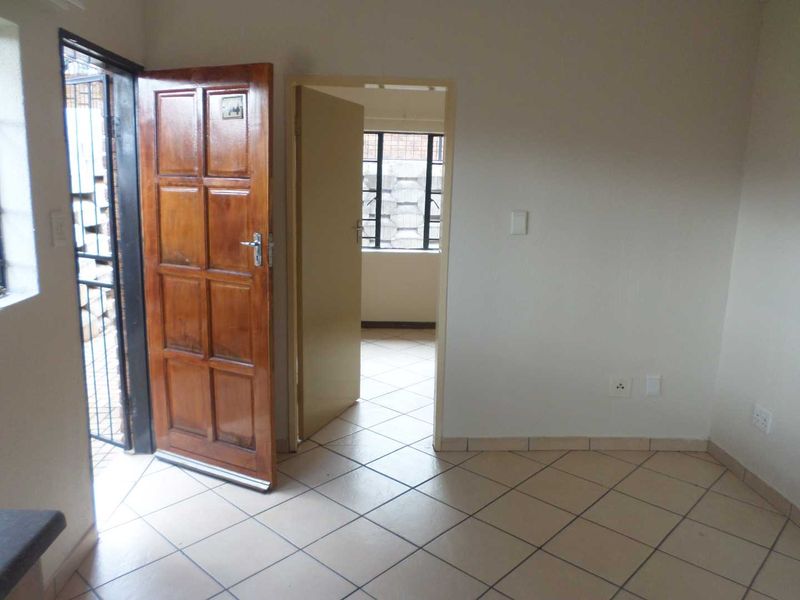 An empty living area featuring tiled flooring, a timber entrance door with security gate, and direct access to an adjacent room.