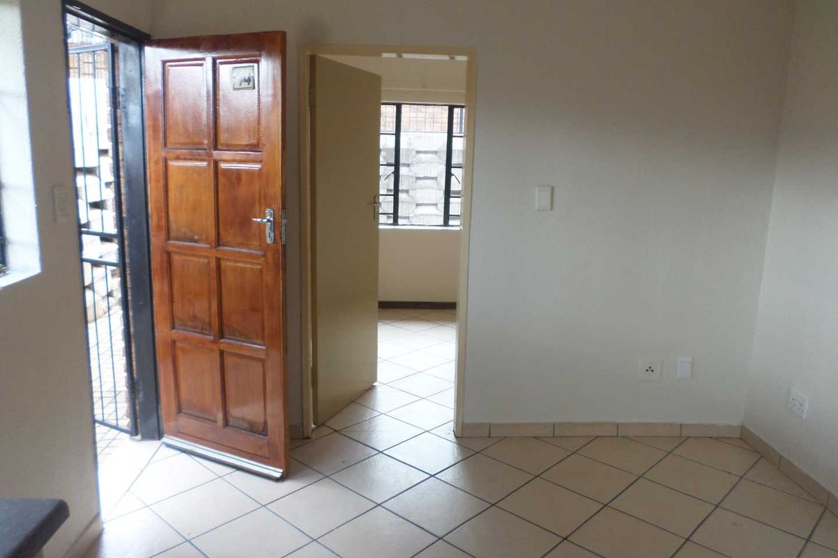 An empty living area featuring tiled flooring, a timber entrance door with security gate, and direct access to an adjacent room.