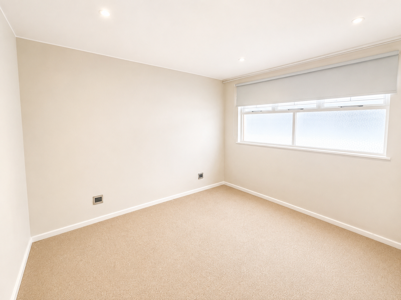 A clean, neutral-toned bedroom featuring carpet flooring, recessed downlighting, and a fitted roller blind over the window.