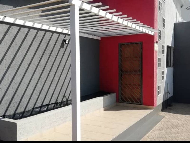 A modern covered patio area featuring a white pergola, built-in planter, and secure entrance door, finished with clean paving and contrasting exterior wall paint.