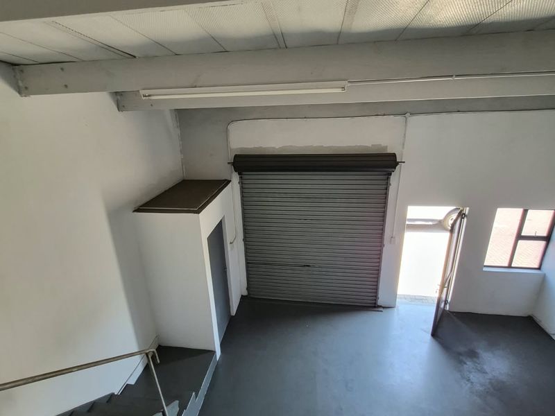 Industrial warehouse space featuring a high roller shutter door, exposed ceiling beams, concrete flooring, and an internal staircase leading to the upper level.