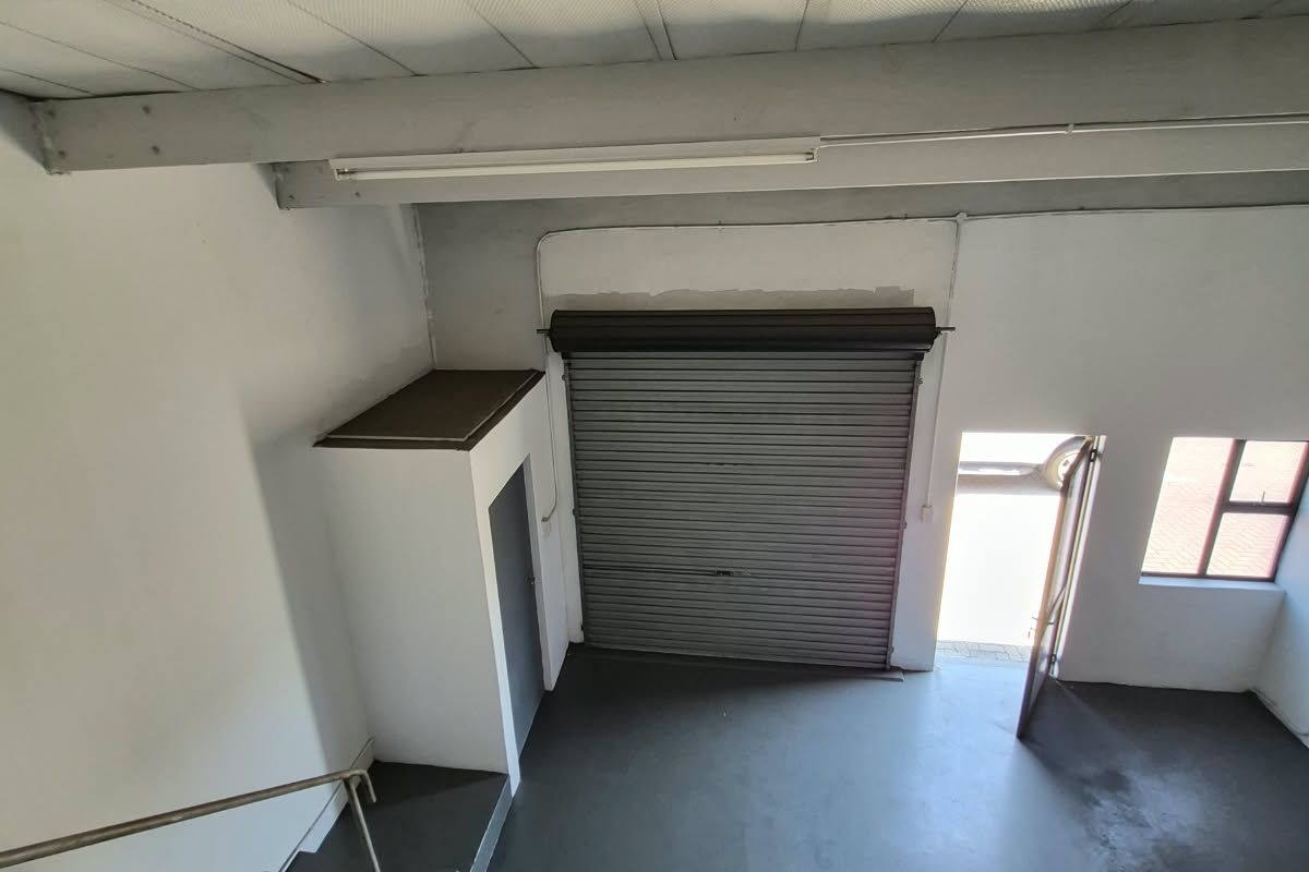 Industrial warehouse space featuring a high roller shutter door, exposed ceiling beams, concrete flooring, and an internal staircase leading to the upper level.
