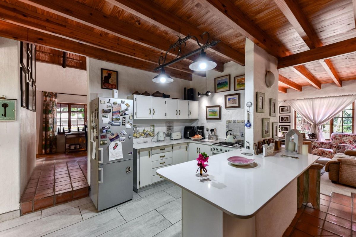 A rustic kitchen featuring exposed timber ceiling beams, a central island, white cabinetry, and tiled flooring, seamlessly connecting to adjacent living spaces.