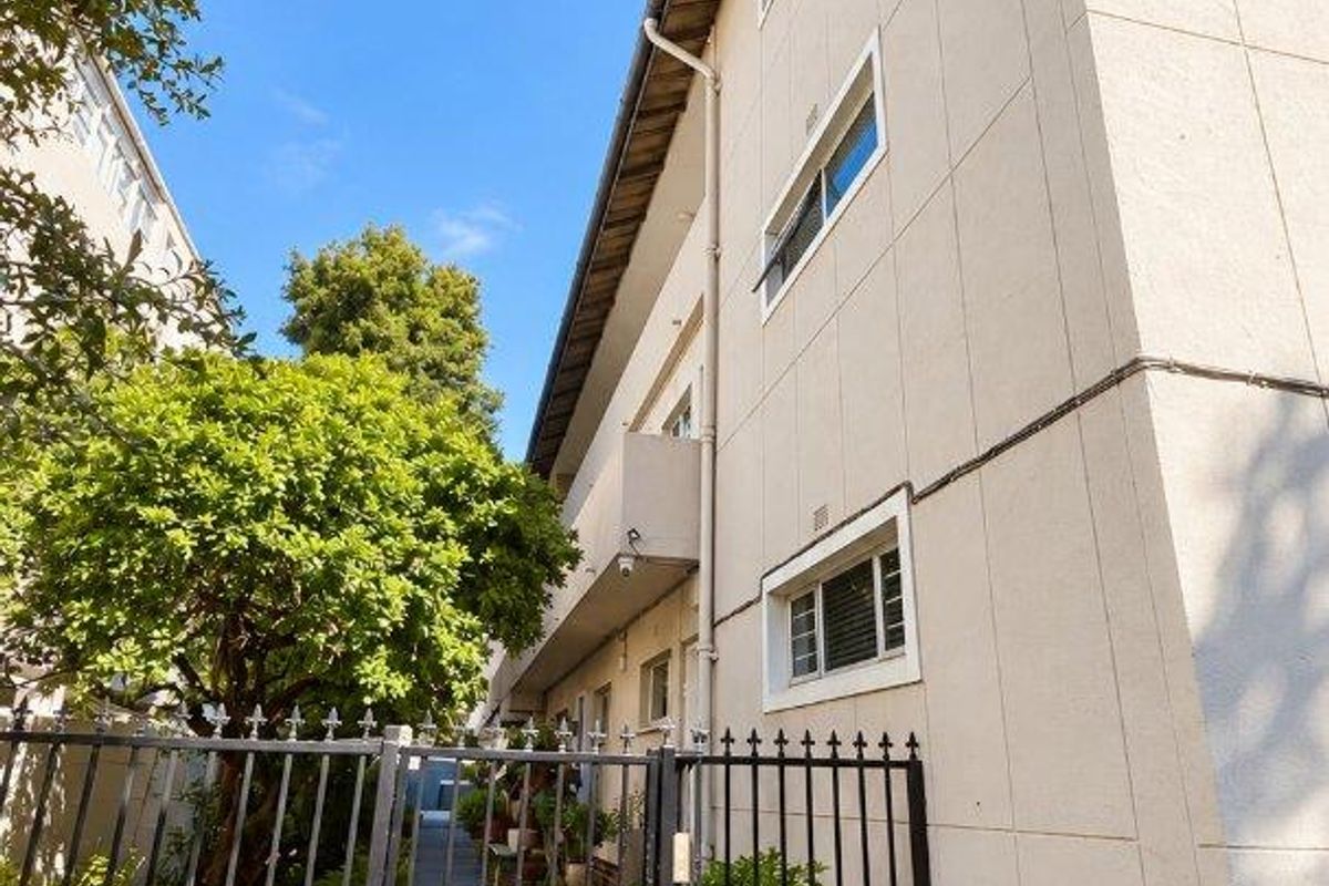 A view of the apartment building's rear exterior, featuring a paved walkway, wrought iron security gate, and multi-level facade with balconies.