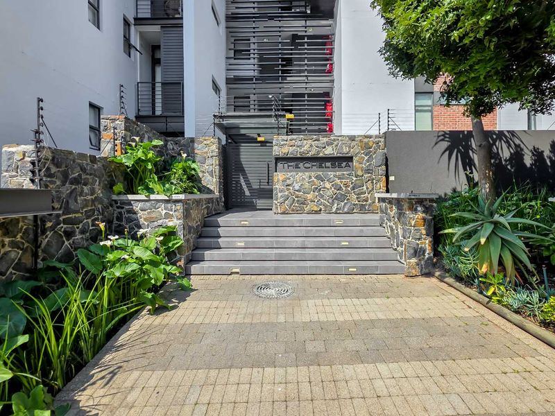 The front entrance of The Chelsea apartment complex features a stone-clad boundary wall, paved driveway, and secure gated access with integrated step lighting.
