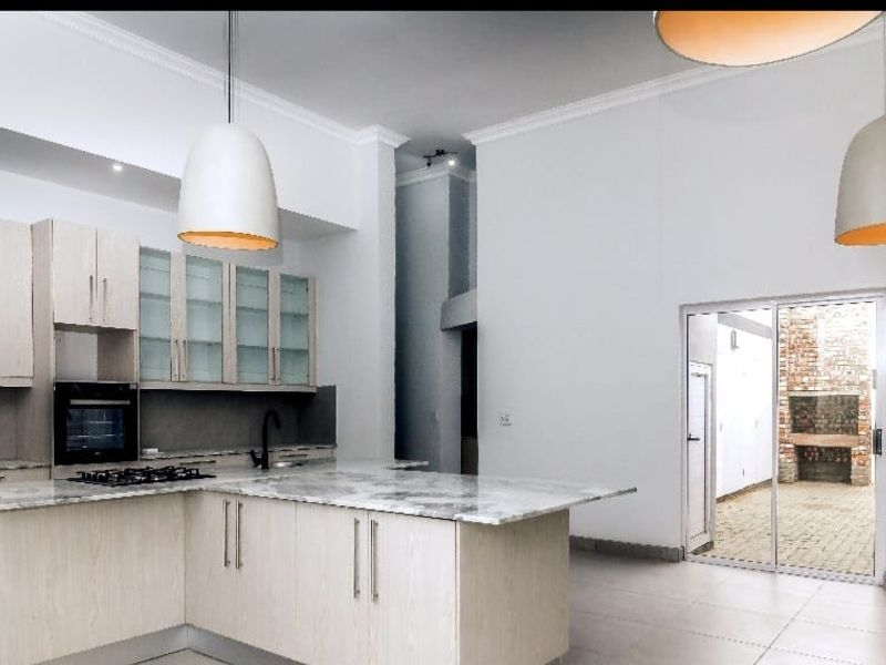 Spacious open-plan kitchen featuring light wood cabinetry, stone countertops, and integrated appliances, flowing seamlessly to an outdoor patio area with a built-in brick braai.