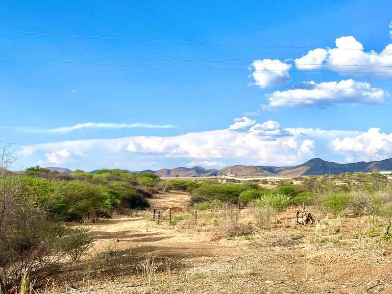 A wide-angle view of the vacant commercial land featuring natural vegetation, a dirt access track, and wire fencing against a mountain backdrop.