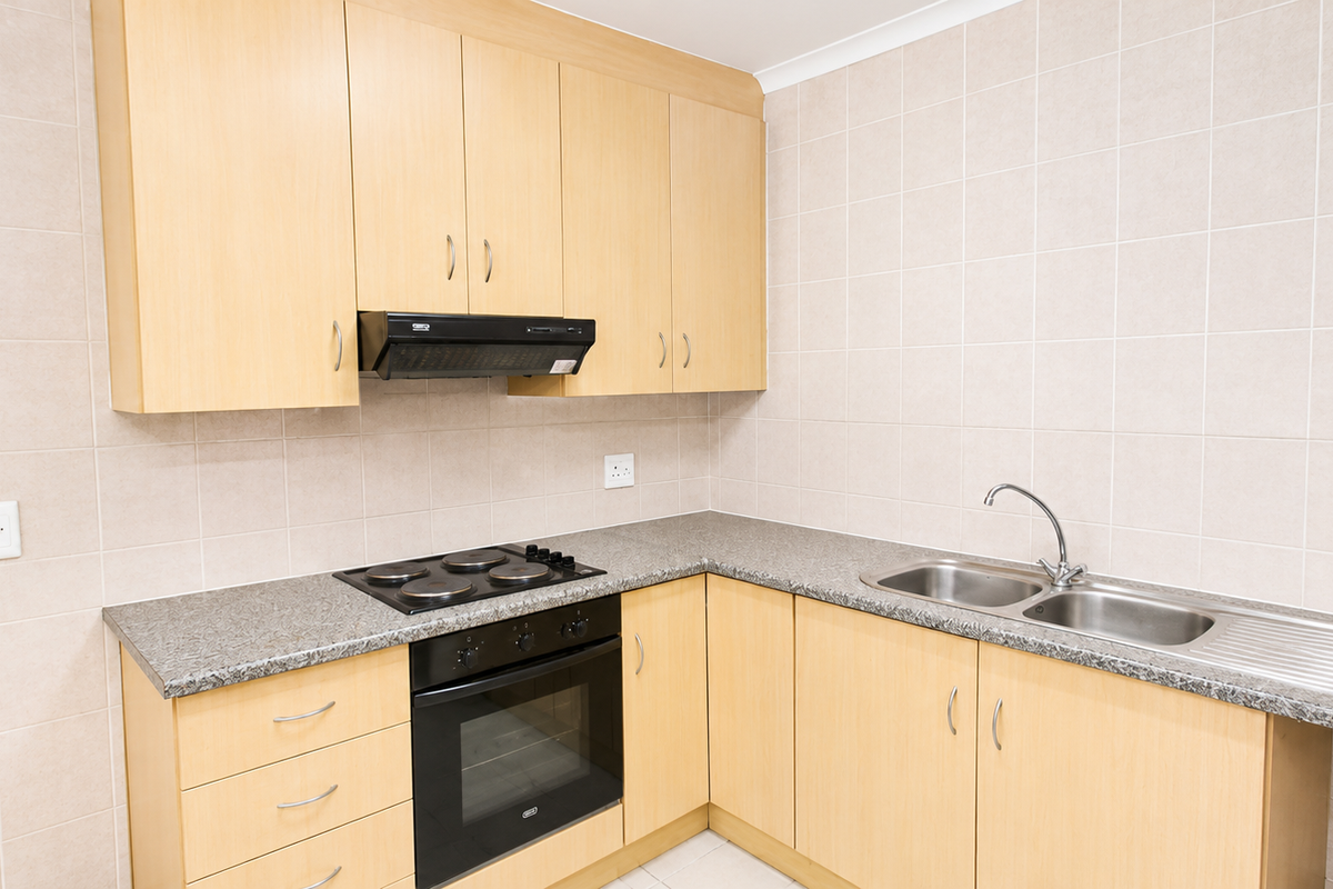 A clean, functional kitchen featuring light wood-finish cabinetry, granite countertops, a built-in oven, electric hob, and a stainless steel double sink with tiled splashbacks.