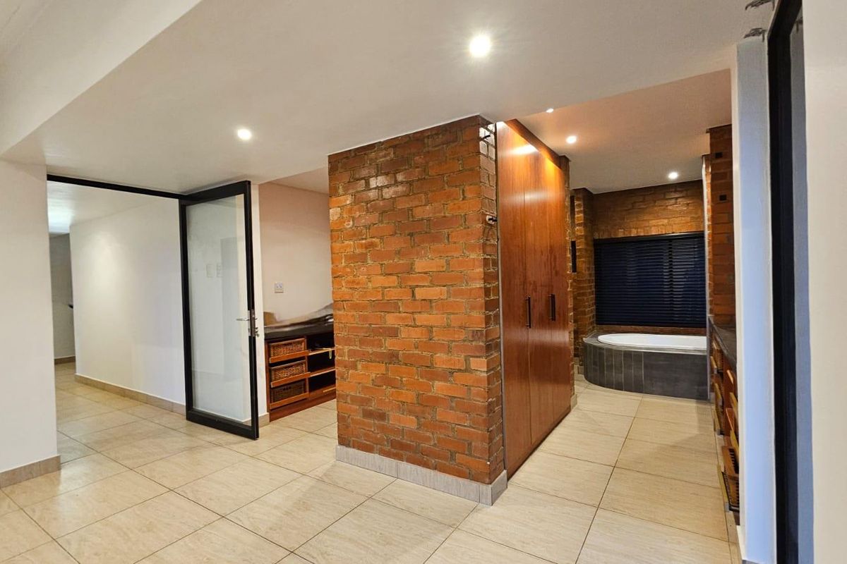 A spacious bathroom featuring a central exposed brick feature wall, built-in wooden cabinetry, recessed lighting, and a corner bathtub.