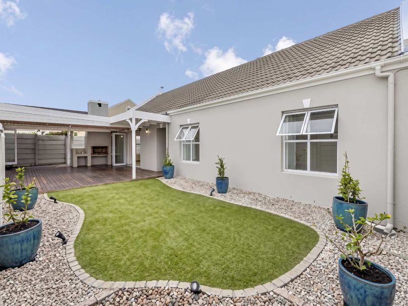 A well-maintained rear exterior featuring a covered patio with built-in braai, timber decking, and a landscaped garden with artificial lawn and pebble borders.