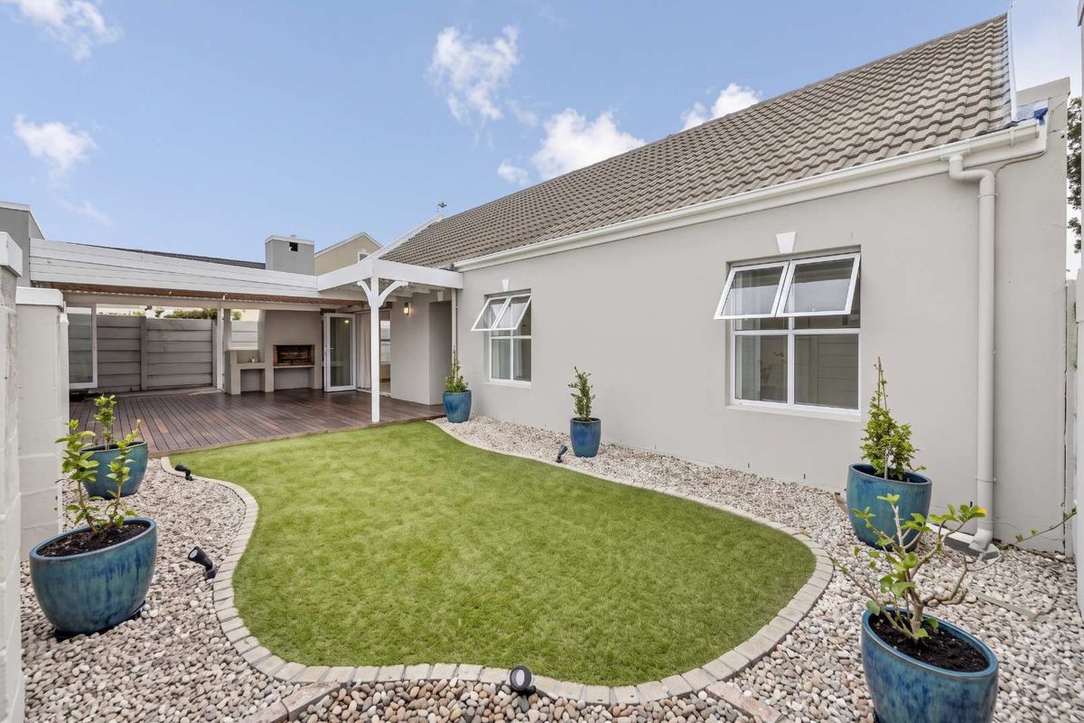 A well-maintained rear exterior featuring a covered patio with built-in braai, timber decking, and a landscaped garden with artificial lawn and pebble borders.