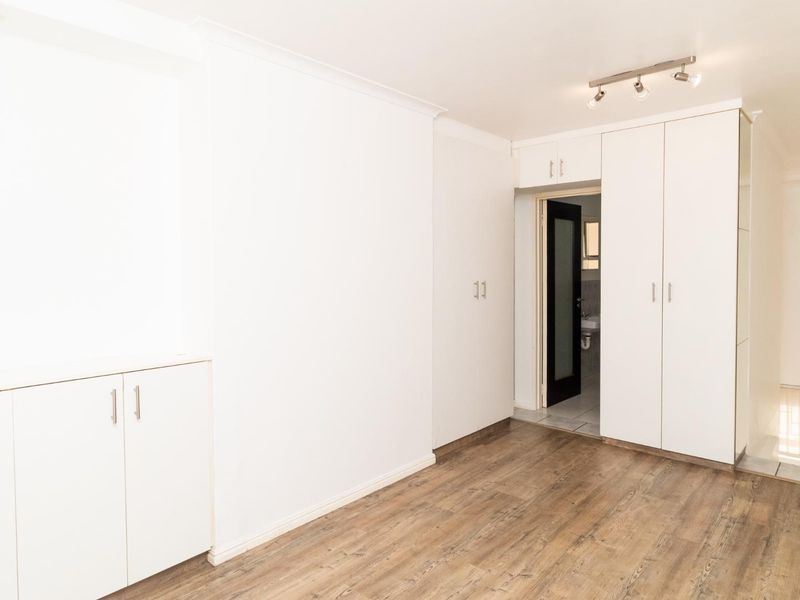 A bright, empty bedroom featuring modern laminate flooring, extensive built-in cupboard storage, and clean white finishes throughout.