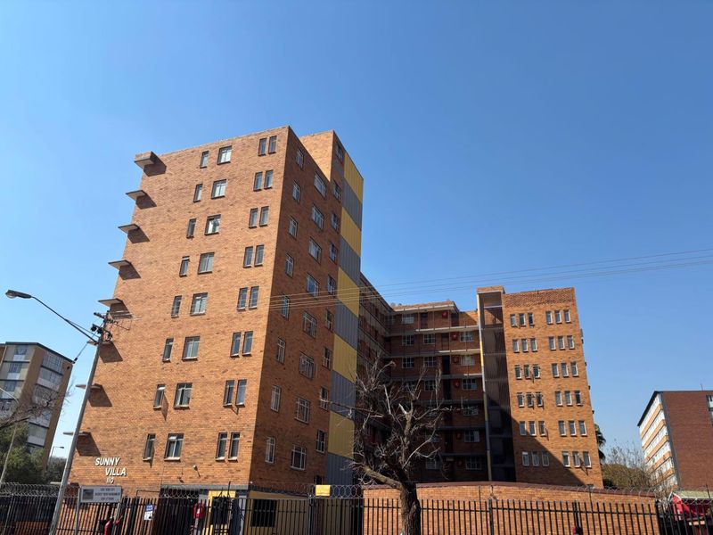 Exterior view of Sunny Villa apartment complex featuring a multi-story brick facade, secure perimeter fencing, and clear street-level access.