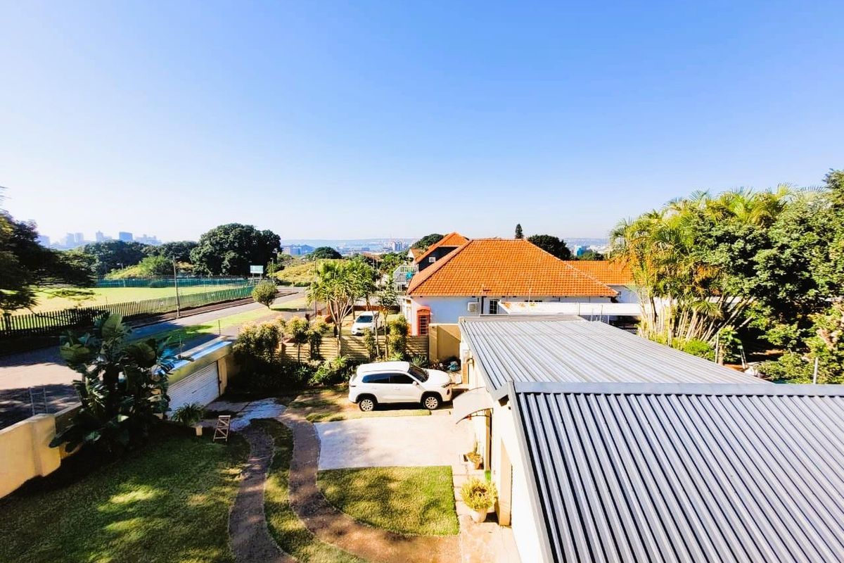 Elevated view overlooking the property grounds, featuring a metal roof, landscaped garden, paved driveway, and surrounding neighbourhood scenery.