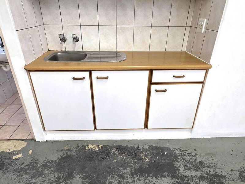 A compact kitchenette area featuring a stainless steel sink, laminate countertop, and tiled backsplash, adjacent to a small tiled washroom area.