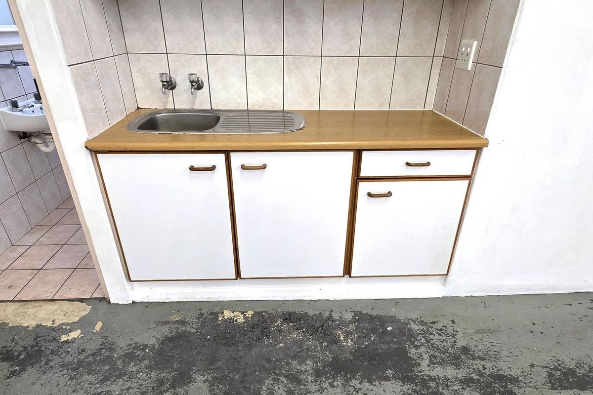 A compact kitchenette area featuring a stainless steel sink, laminate countertop, and tiled backsplash, adjacent to a small tiled washroom area.