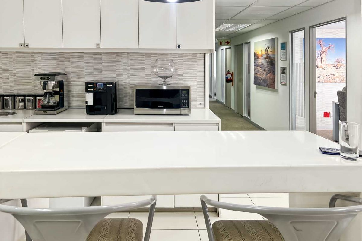 A clean office kitchenette area featuring white cabinetry, a tiled backsplash, and a built-in counter, leading into a hallway with multiple office doors.